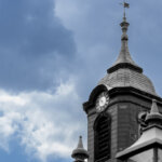 Alter Glockenturm mit Laternen vor blauem Himmel mit weißen Wolken